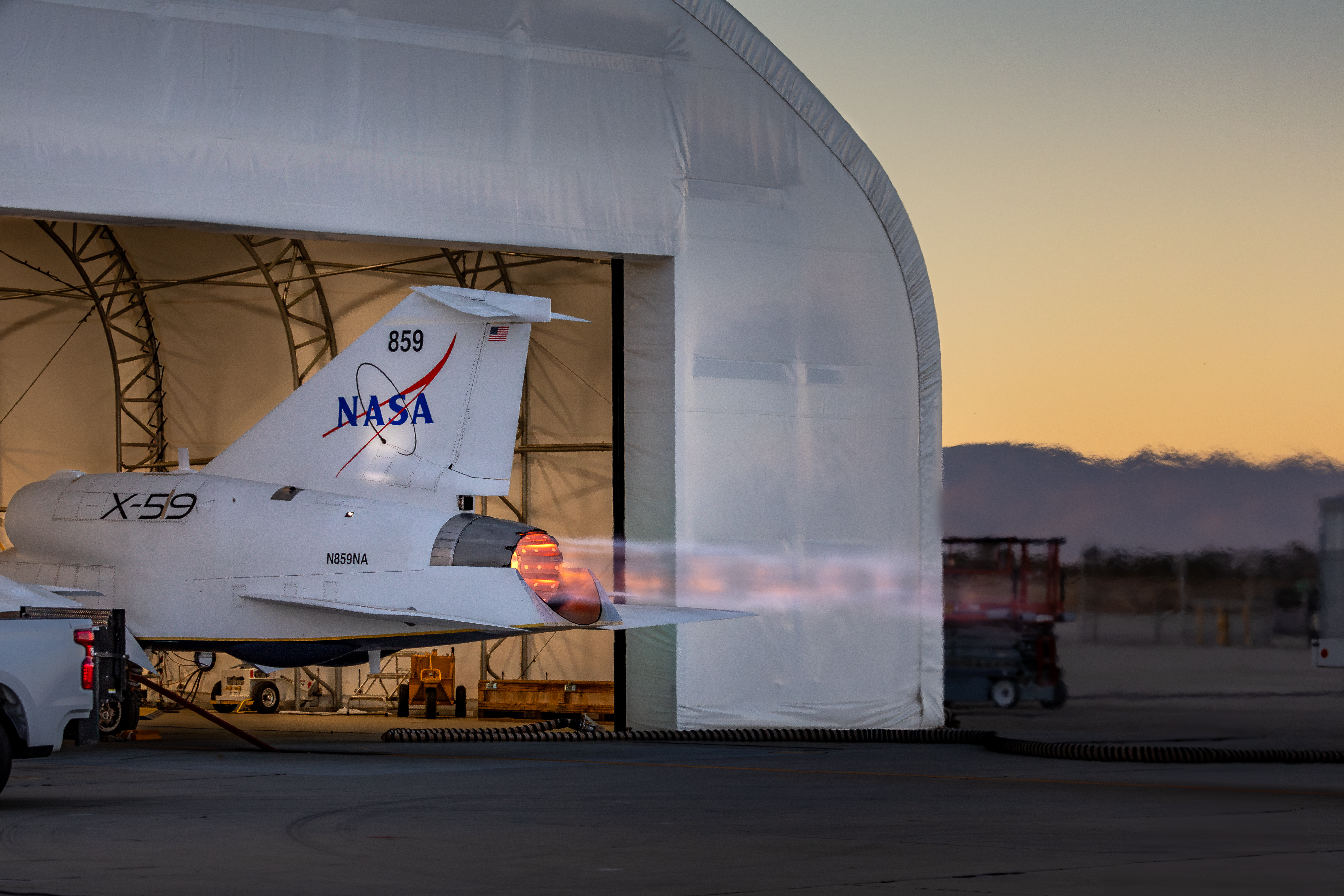 NASA’s X-59 sits partially in a white hangar and emits bright flames with visible shock diamonds from its engine against an evening sky during ma...