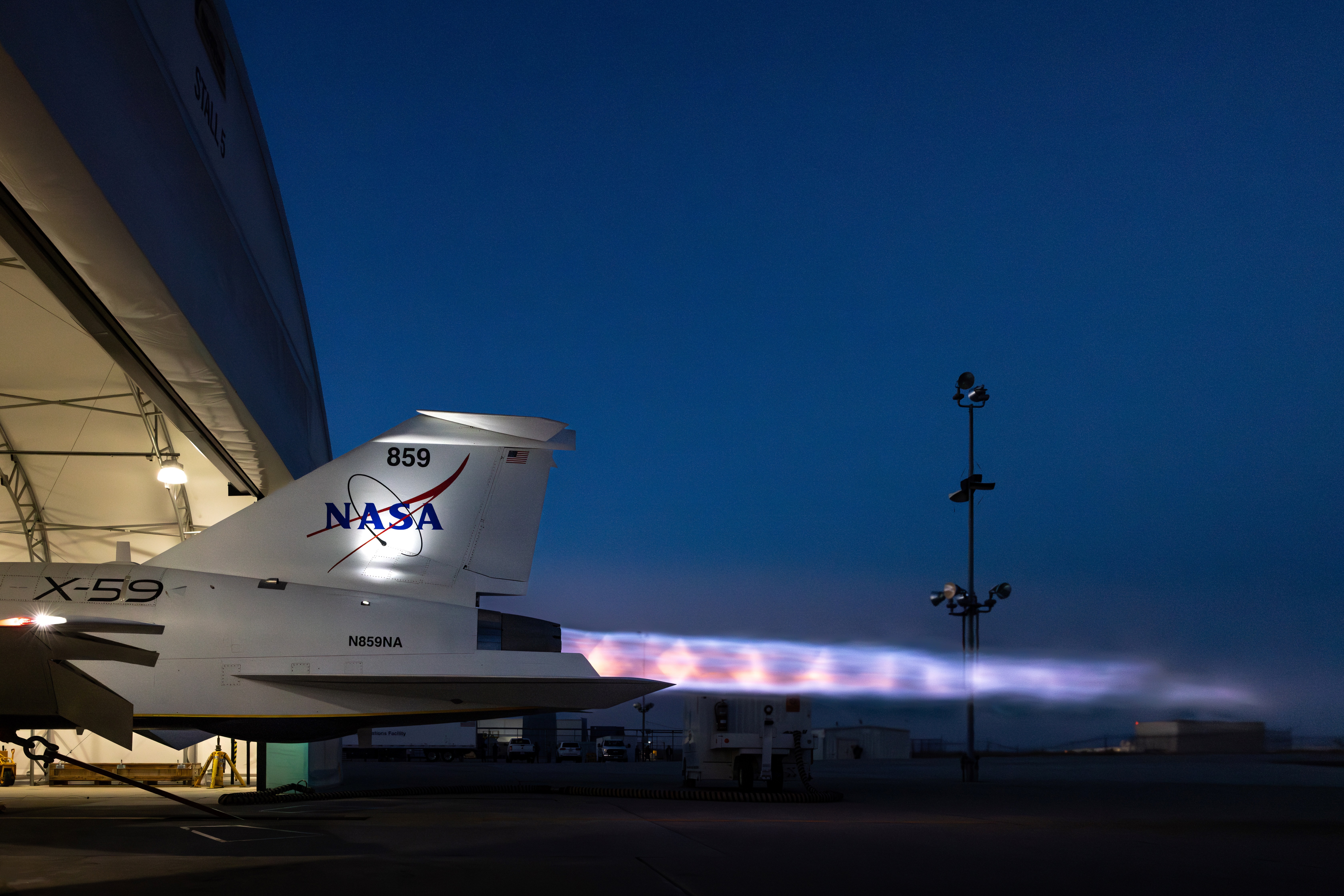 NASA’s X-59 sits partially in a white hangar and emits bright flames with visible shock diamonds from its engine at sunset during maximum afterbu...